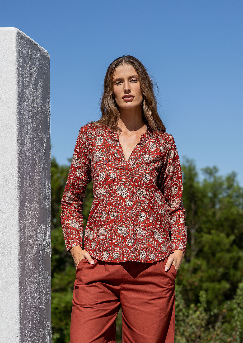 Woman wearing a red floral top and rust-colored pants standing outdoors with trees and blue sky in the background.