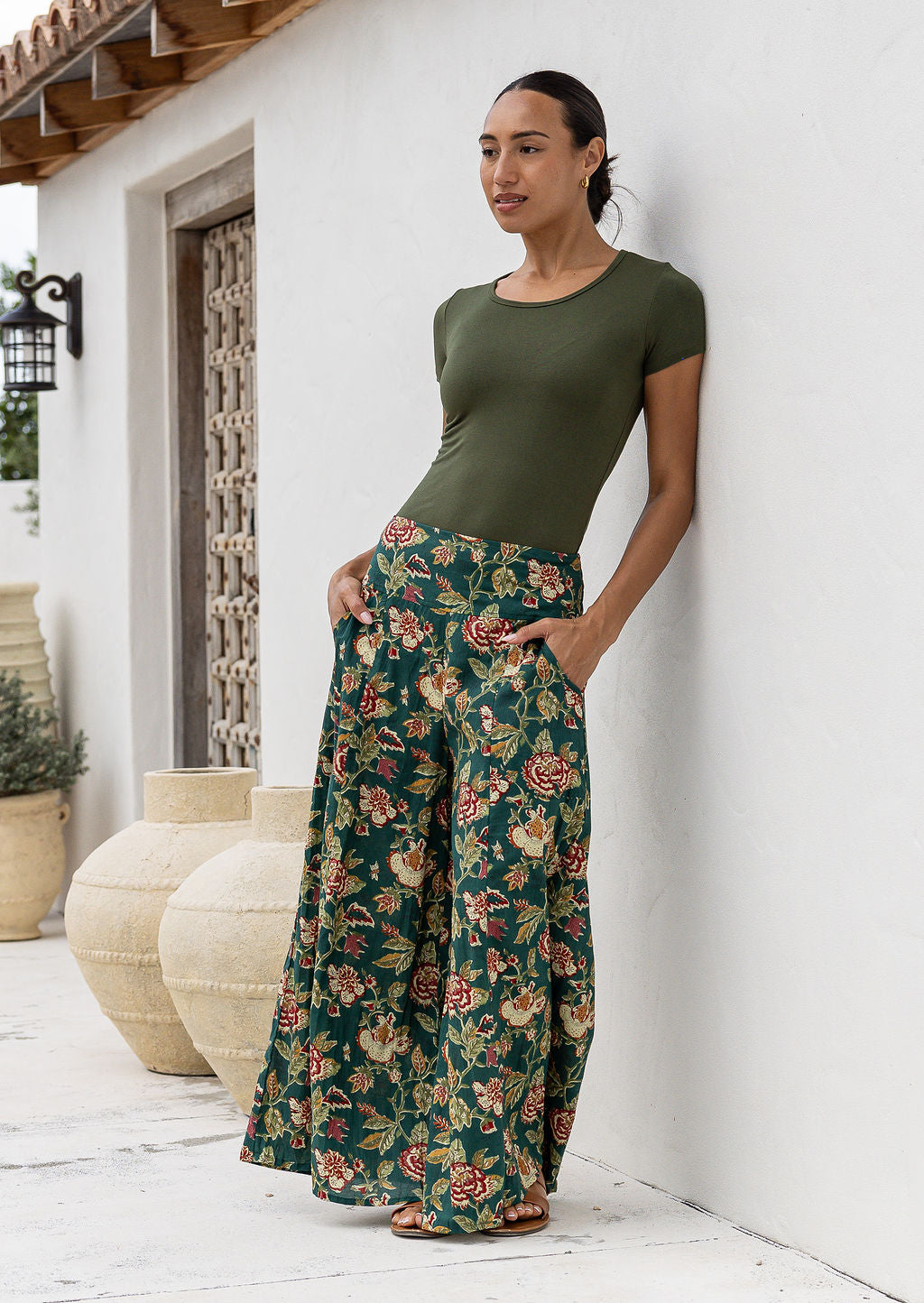 Woman wearing a green top and wide legged green cotton floral pants standing against a white wall with hands in pockets