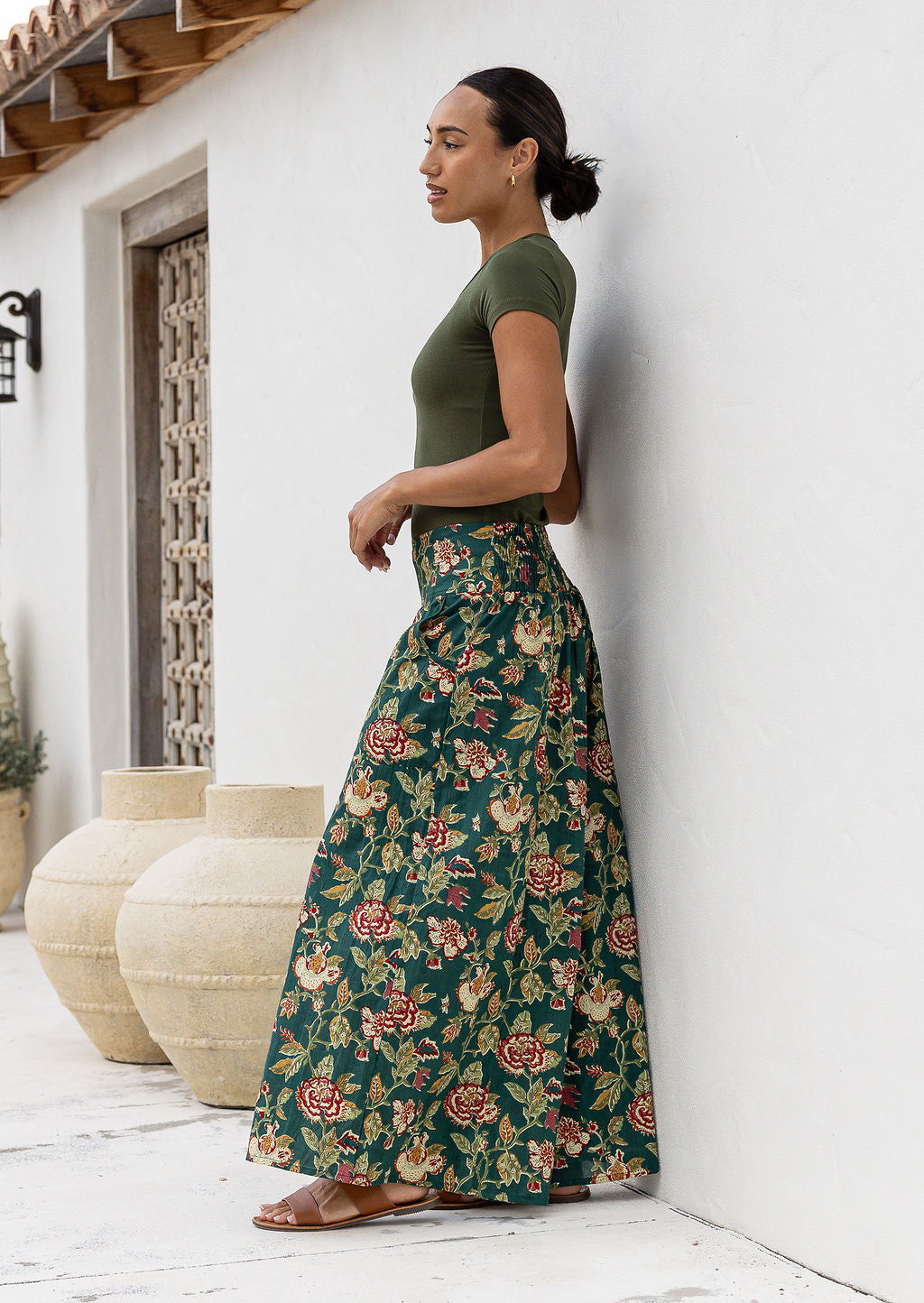 Woman wearing a green t-shirt and wide legged green cotton floral pants standing against a white wall.