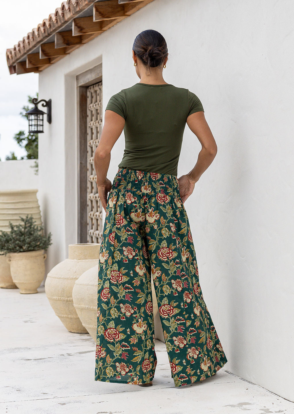 Woman wearing a green top and wide legged green cotton floral pants standing against a white wall back view of hands in pockets