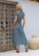 Woman standing in a back pose wearing a 100% rayon wrap dress in blue based floral print with butterfly sleeves and long fabric ties