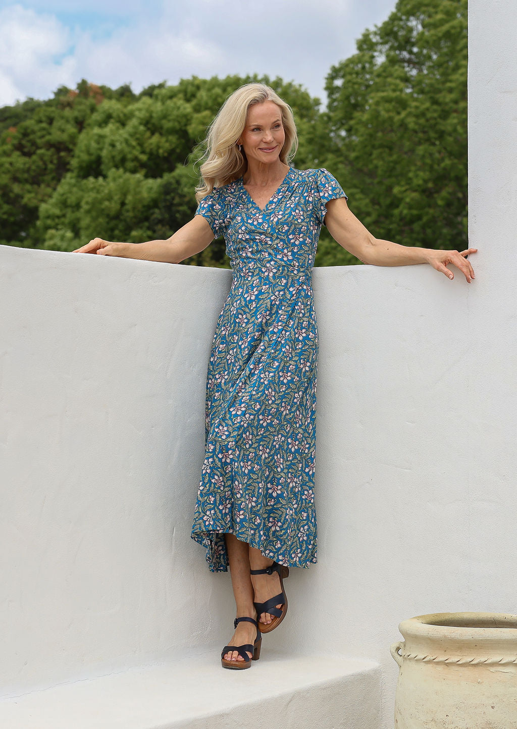 Woman wearing a blue floral wrap style maxi dress stands on a white concrete surface with trees in the background