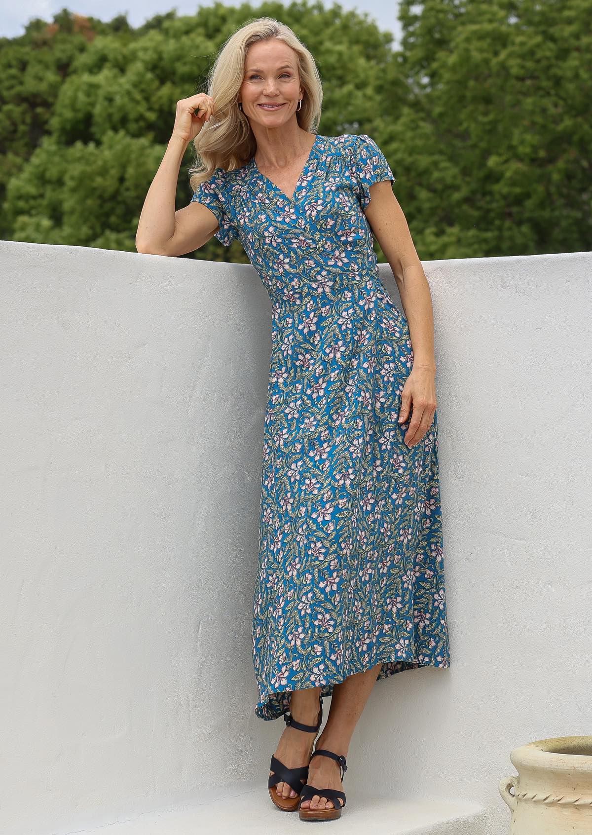 Woman in a blue floral rayon maxi dress with wrap around style and V-neckline, standing on a white ledge with green background