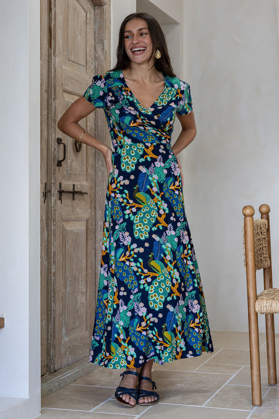 Woman wearing a navy blue based floral print wrap around dress with butterfly sleeves, standing in front of a wooden door