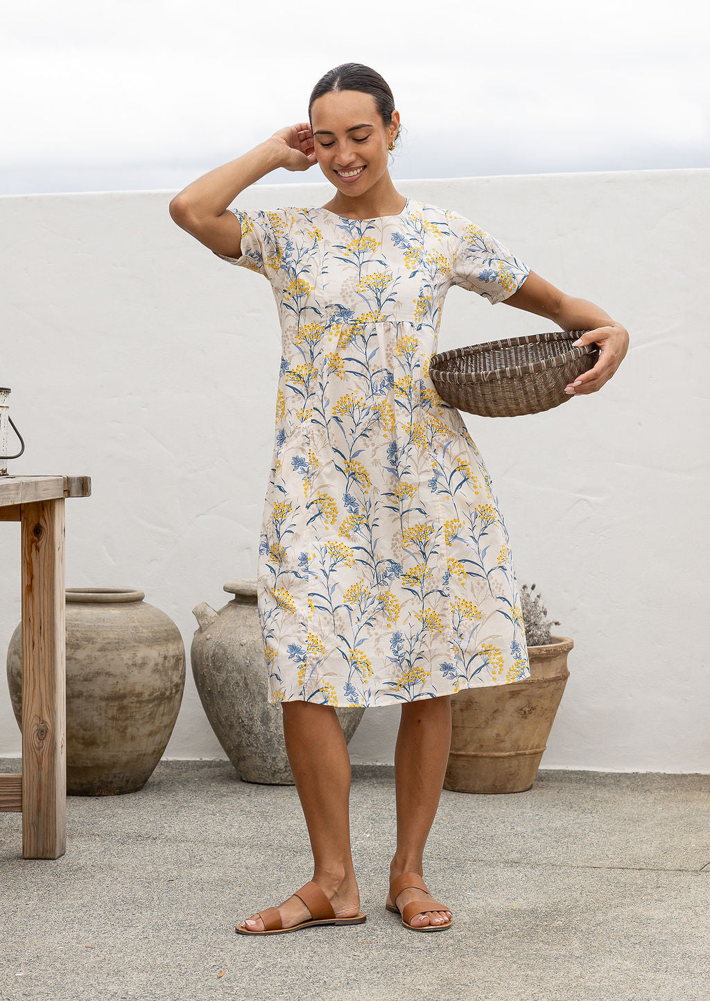 Woman in a 100% cotton floral dress holding a woven basket in an outdoor setting.