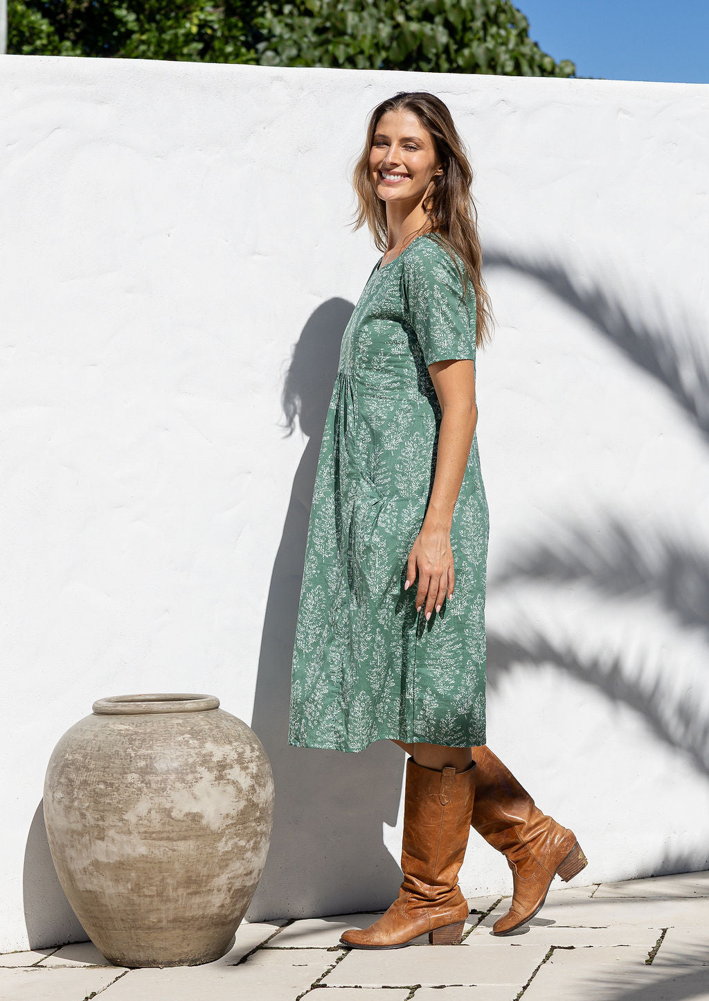 Woman stands sideways wearing a 100% cotton dress in sage green based floral print with short sleeves