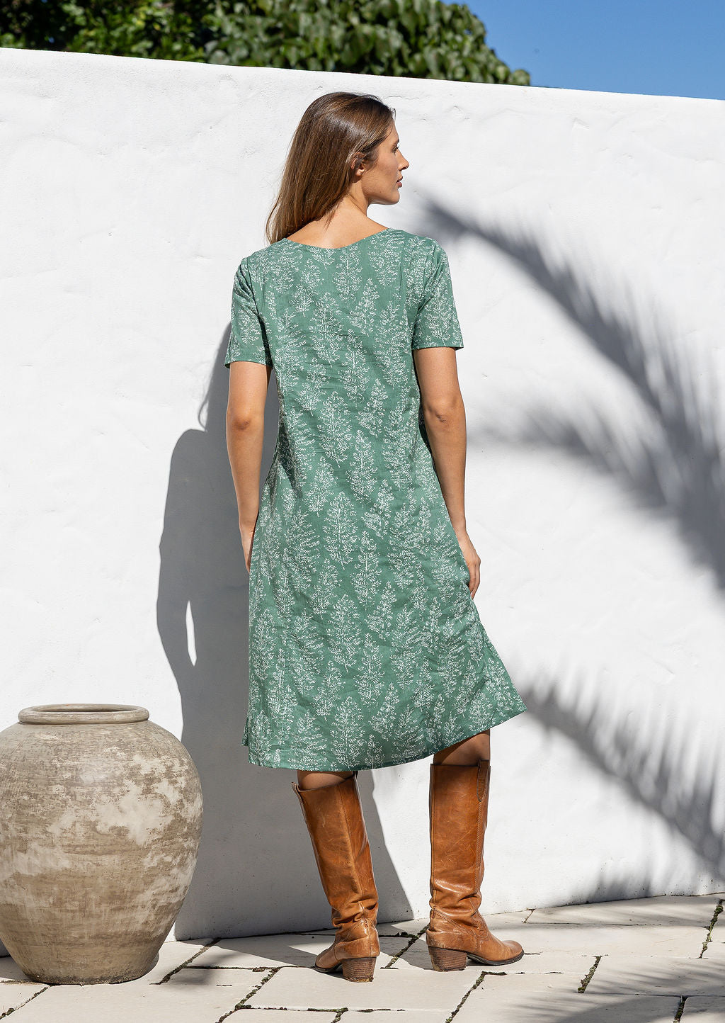 Woman stands backwards wearing a 100% cotton dress in sage green based floral print with short sleeves and brown boots