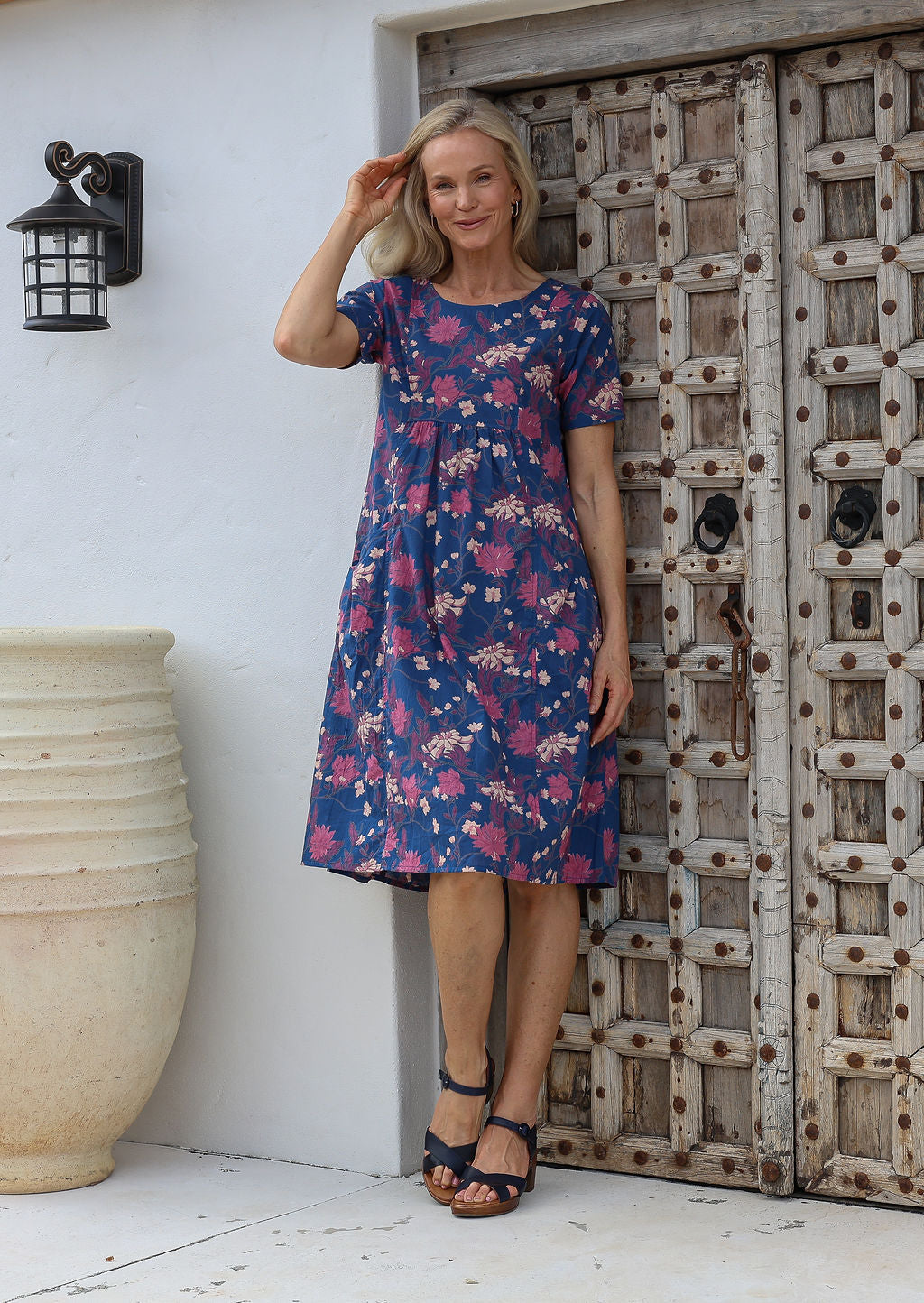 Woman standing in front of a rustic wooden door wears 100% cotton below knee length loose fit dress with short sleeves and round neckline.