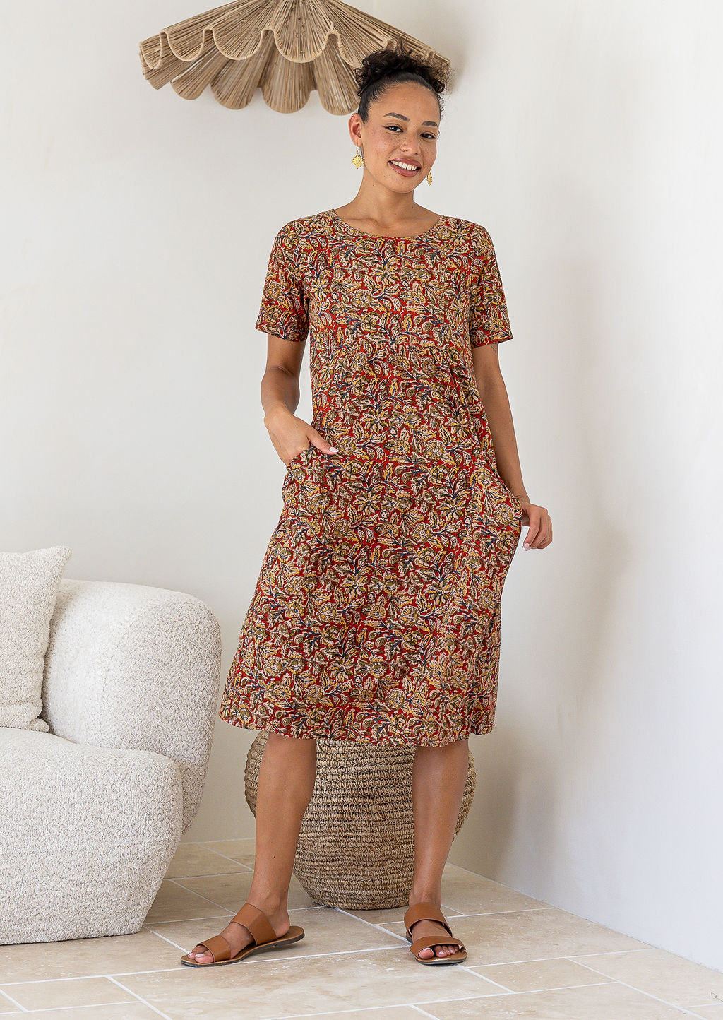 Woman wearing a patterned floral cotton dress with her hands in the pockets standing in a room with a couch and decorative items.
