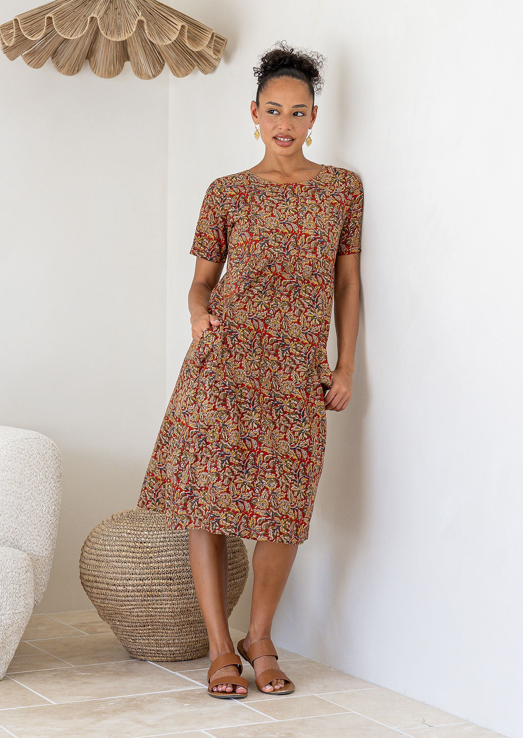 Woman wearing a patterned floral cotton dress with her hand in the pocket standing in a room with a couch and decorative items.