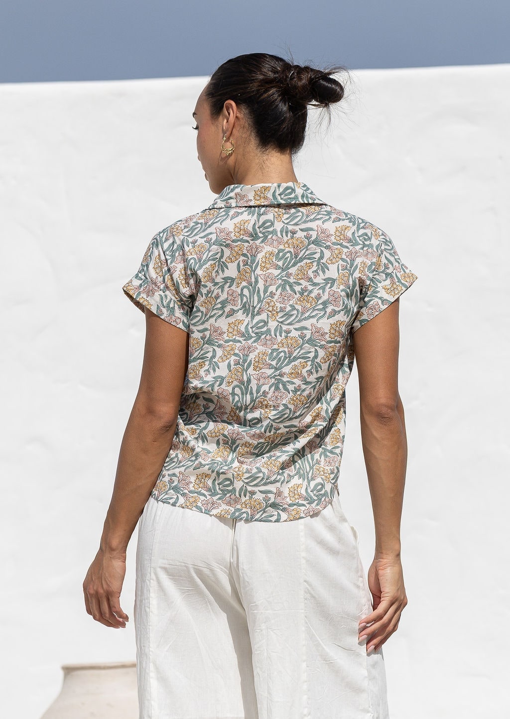 Woman standing backwards wearing a floral cotton top with short folded cuff sleeves, collar and V neckline