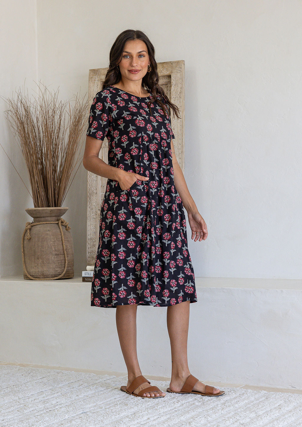 Woman stands sideways wearing a 100% cotton dress in black based floral print with short sleeves and one hand in pocket