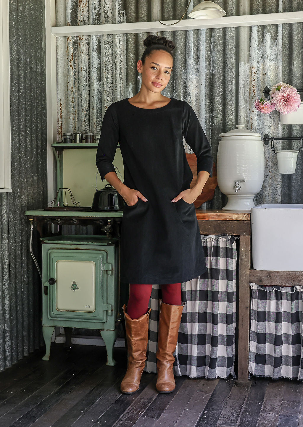 Woman in a black cotton corduroy above knee dress with hands in pockets, standing in a rustic kitchen