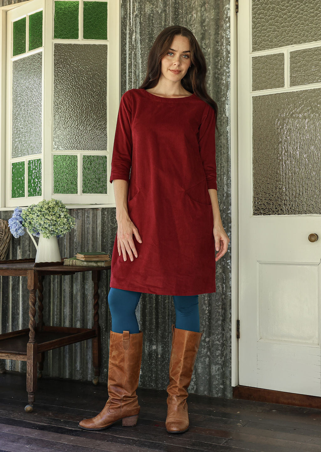 Woman in a red corduroy dress and brown knee-high boots standing in a room with patterned walls.