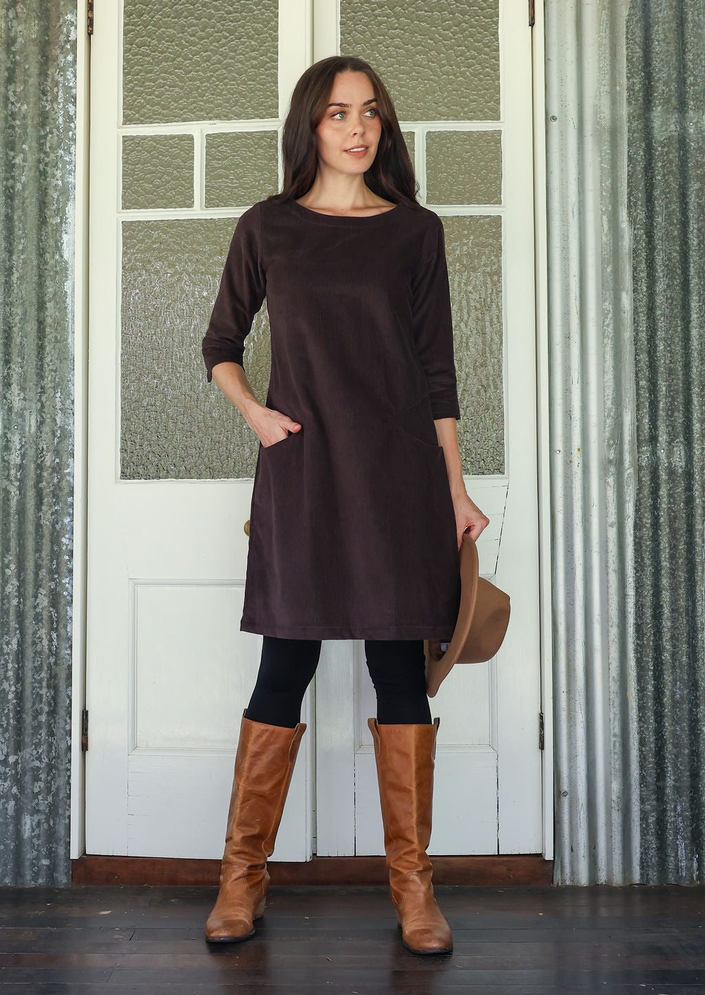 Woman wearing a dark brown dress with long sleeves and brown knee-high boots, standing in front of a white door.