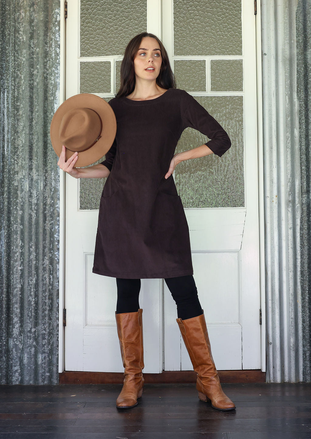 Woman in a brown dress and boots holding a hat against a textured wall.