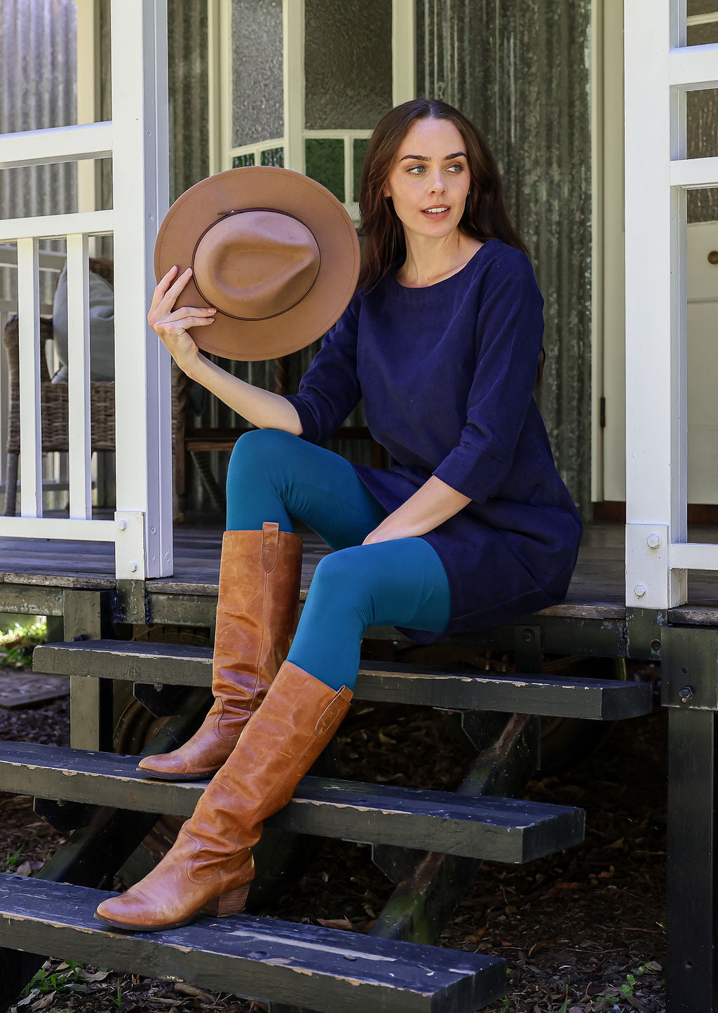 Woman sitting on steps wearing a blue dress, blue leggings, and brown knee-high boots, holding a brown hat.