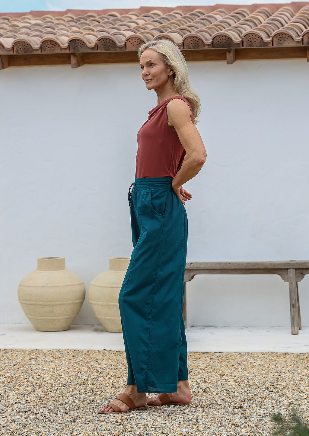 Woman wearing a rust top and deep teal 100% cotton, wide-legged pants standing outdoors with a white wall and wooden bench in the background.