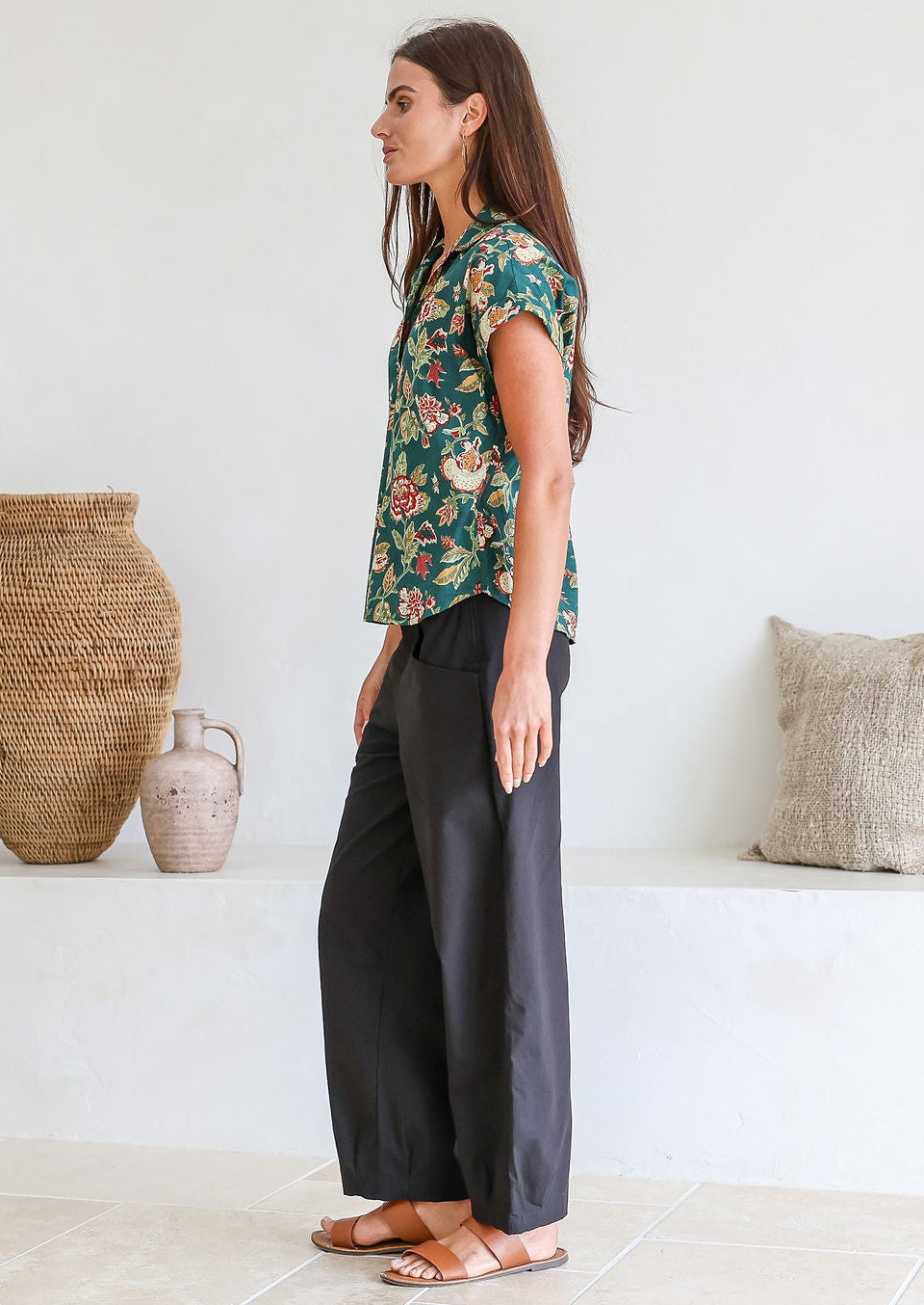 Model stands sideways wearing a 100 percent cotton relaxed fit bottle green based floral top with short folded cuff sleeves, collar and V neckline