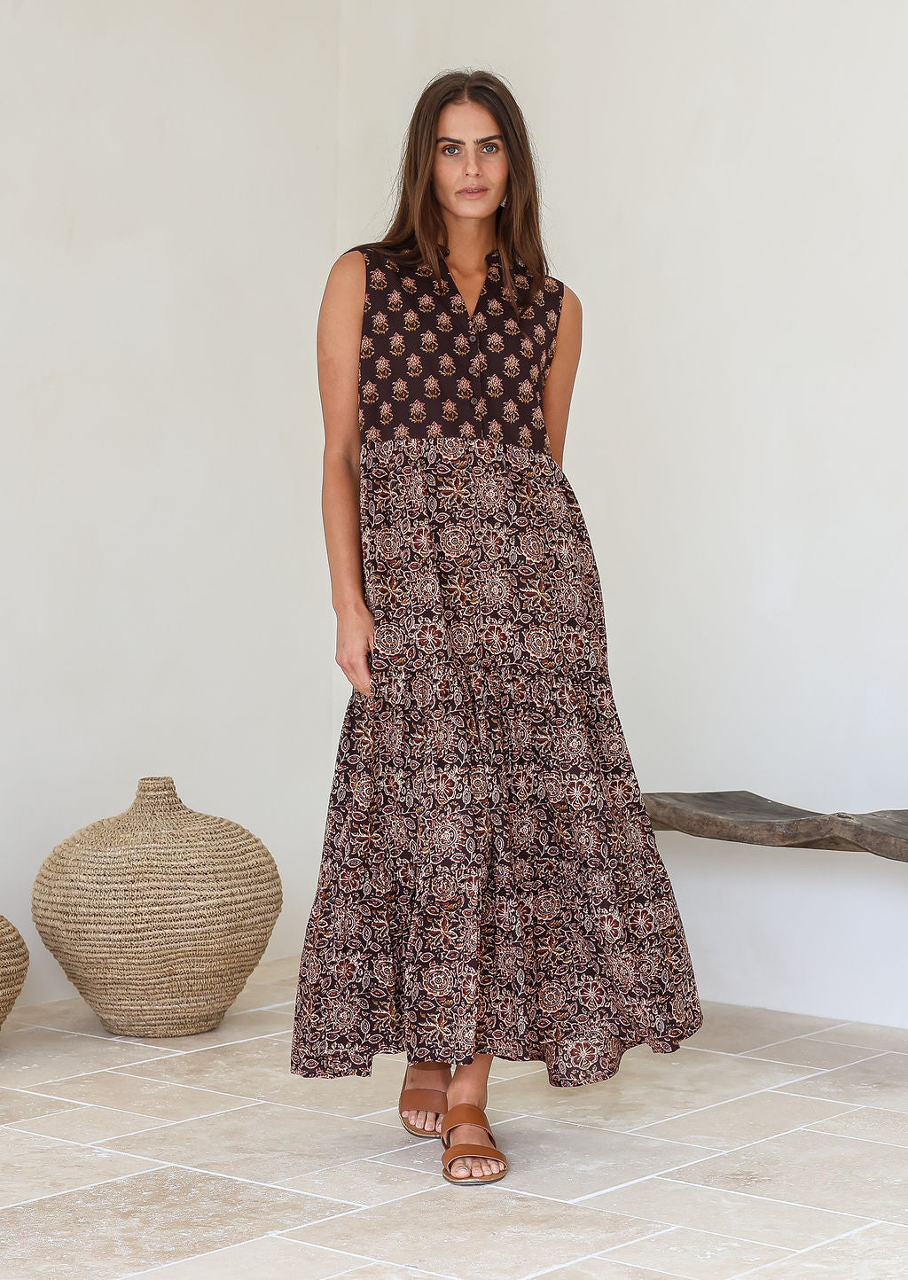 Woman wearing a brown printed sleeveless floral cotton dress standing in a minimalistic room with a woven basket and wooden bench.