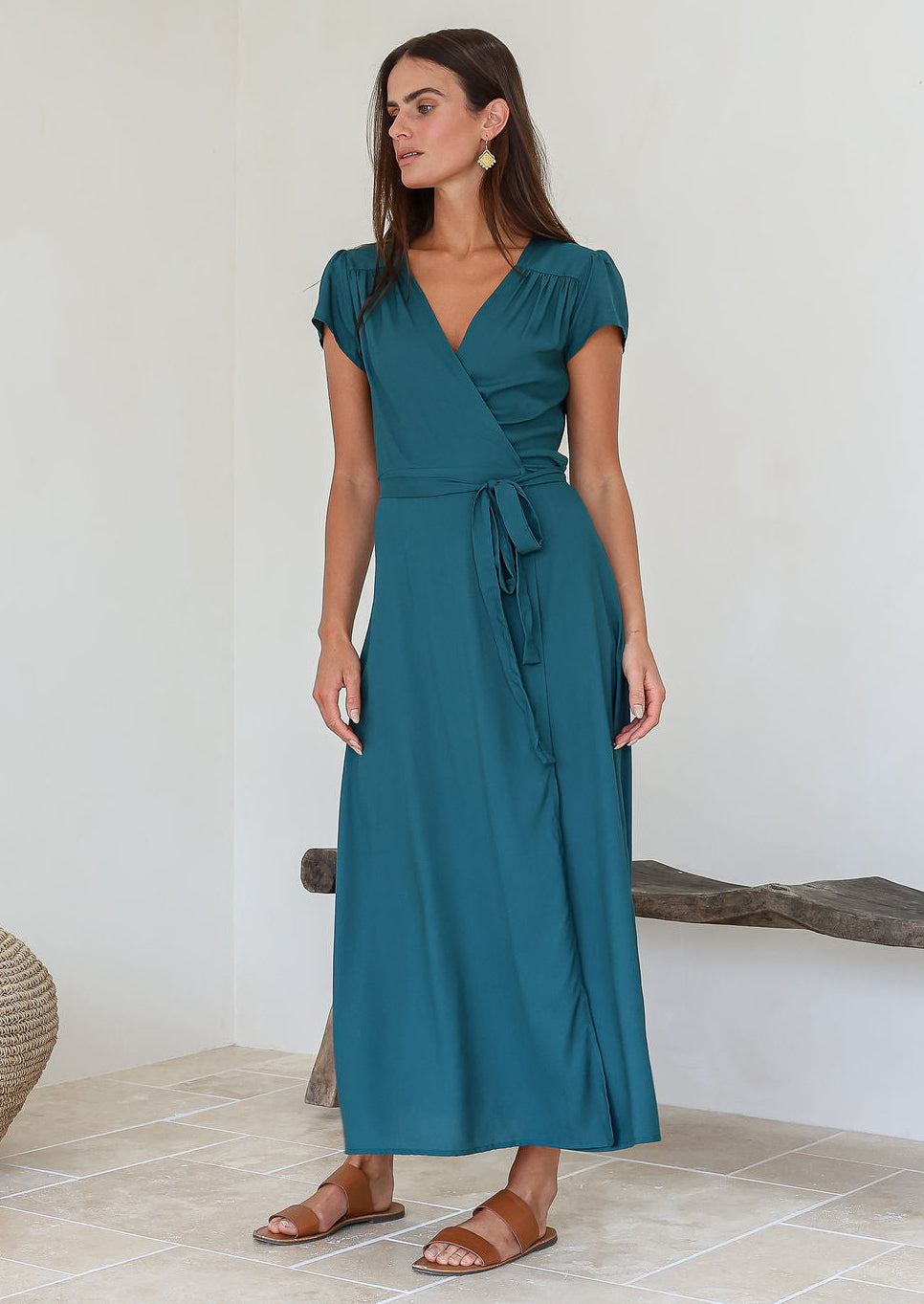 Woman wearing a teal rayon wrap maxi dress standing in a minimal indoor setting.