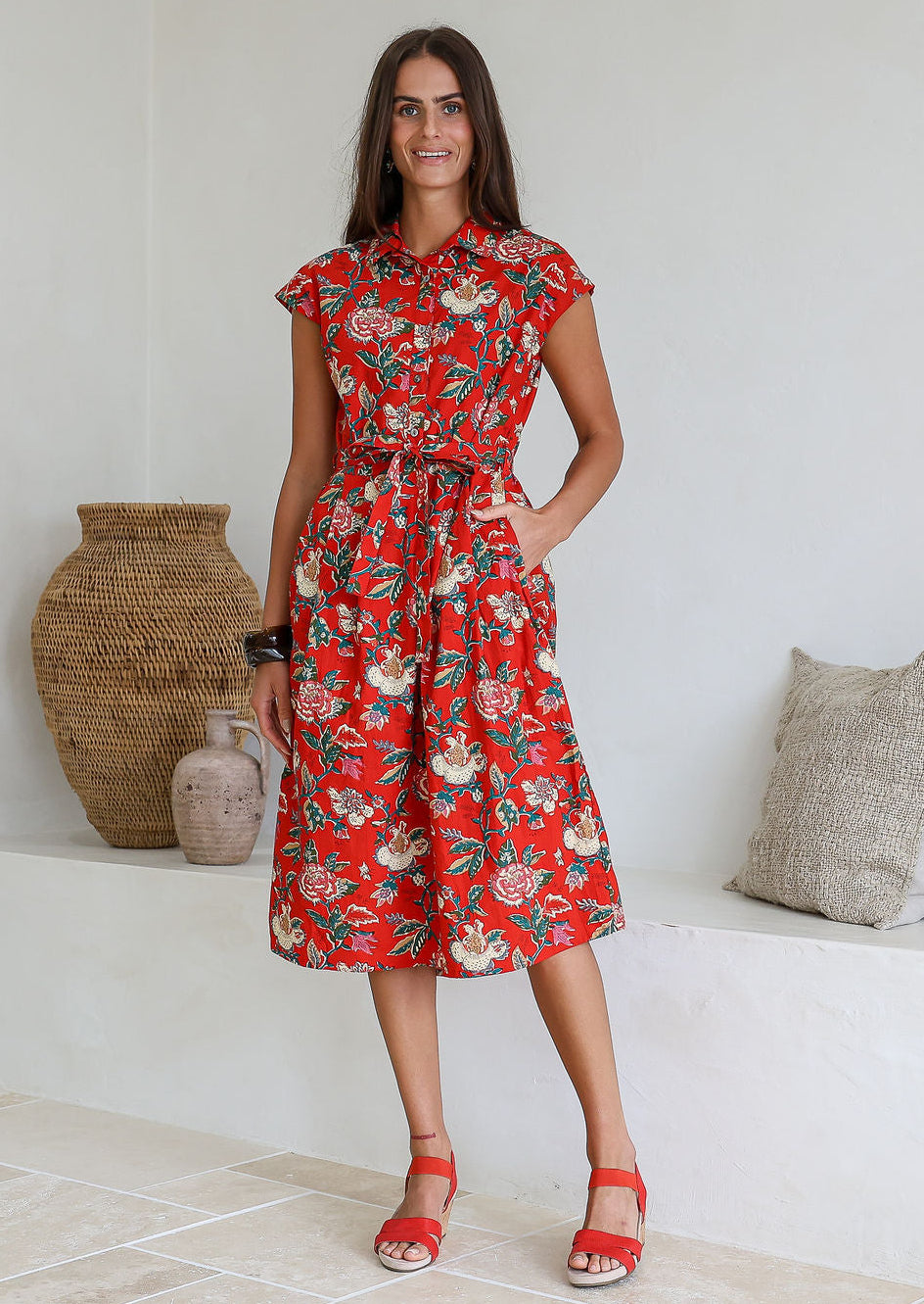 Woman wearing a midi length red based floral cotton dress with buttoned bodice, standing against a white wall and decorative vases