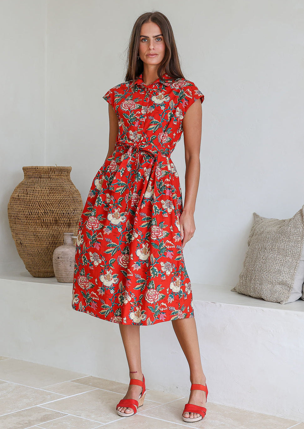 Model stands with wearing a midi length floral printed shirt style cotton dress with cap sleeves, buttoned bodice and a removable waist tie
