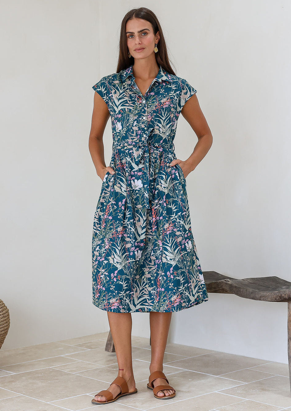 Woman wearing a midi length teal based floral cotton dress with buttoned bodice, standing in a minimal indoor setting.