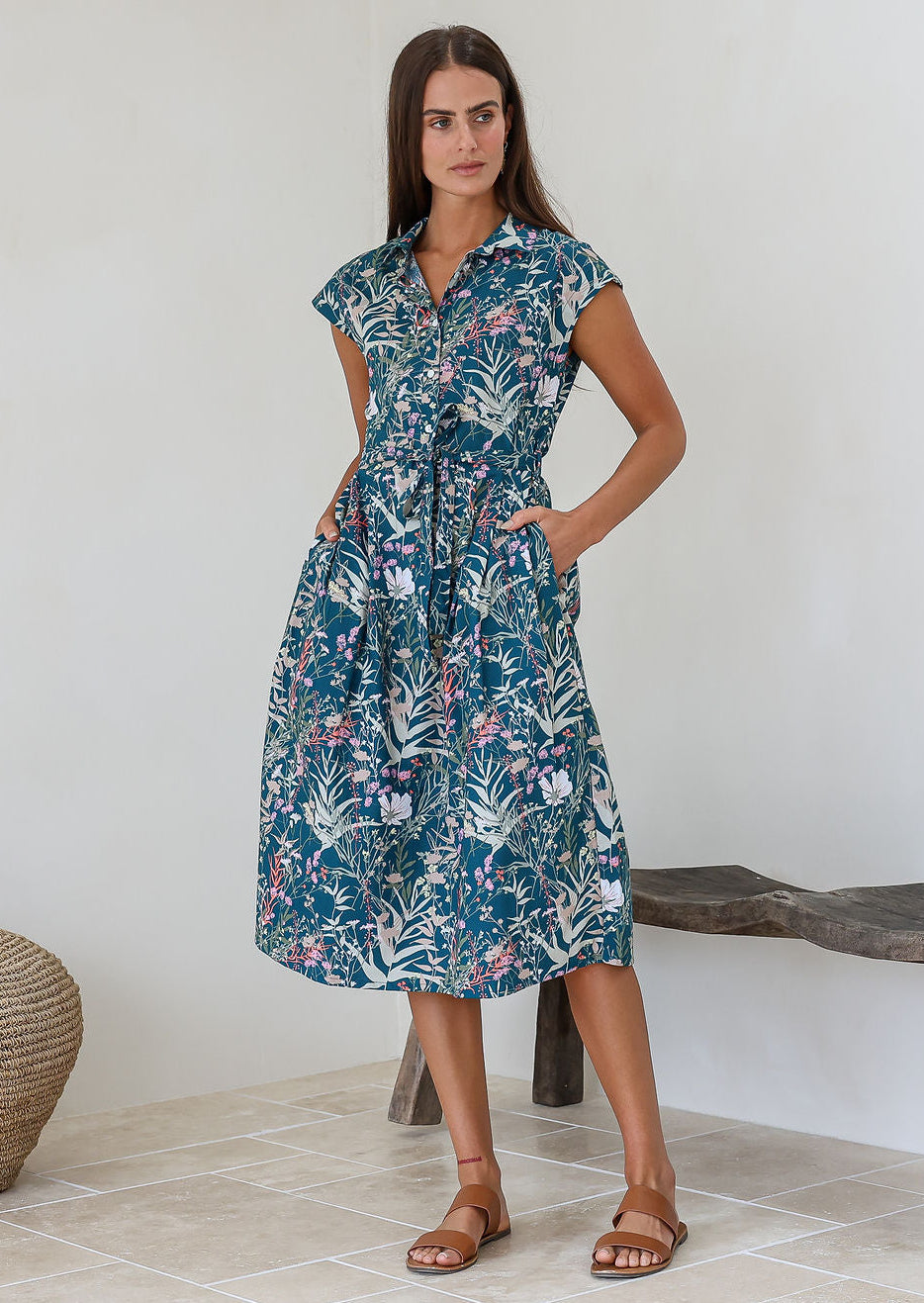 Model stands with both hands in pockets of a midi length floral printed shirt style cotton dress with cap sleeves, buttoned bodice and a removable waist tie