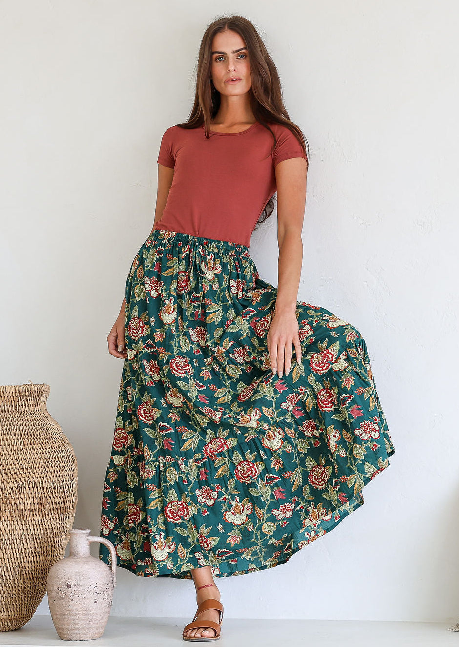 Woman wearing a 100% cotton floral maxi skirt and pink top standing against a white wall with decorative vases.