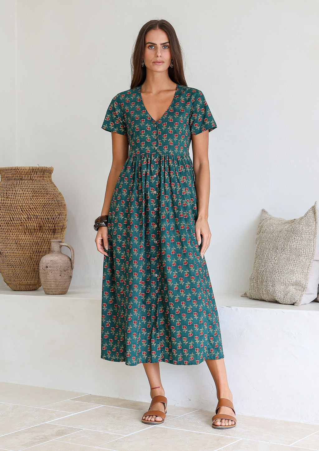 Woman wears 100% cotton button through relaxed fit dress with short sleeves and V-neckline in floral deep red, pink, green and a spot of mustard on a bottle green base.