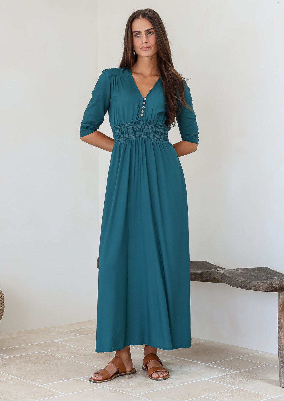 Woman wearing a teal rayon maxi dress with a buttoned bodice and elbow length sleeves, standing against a white wall.