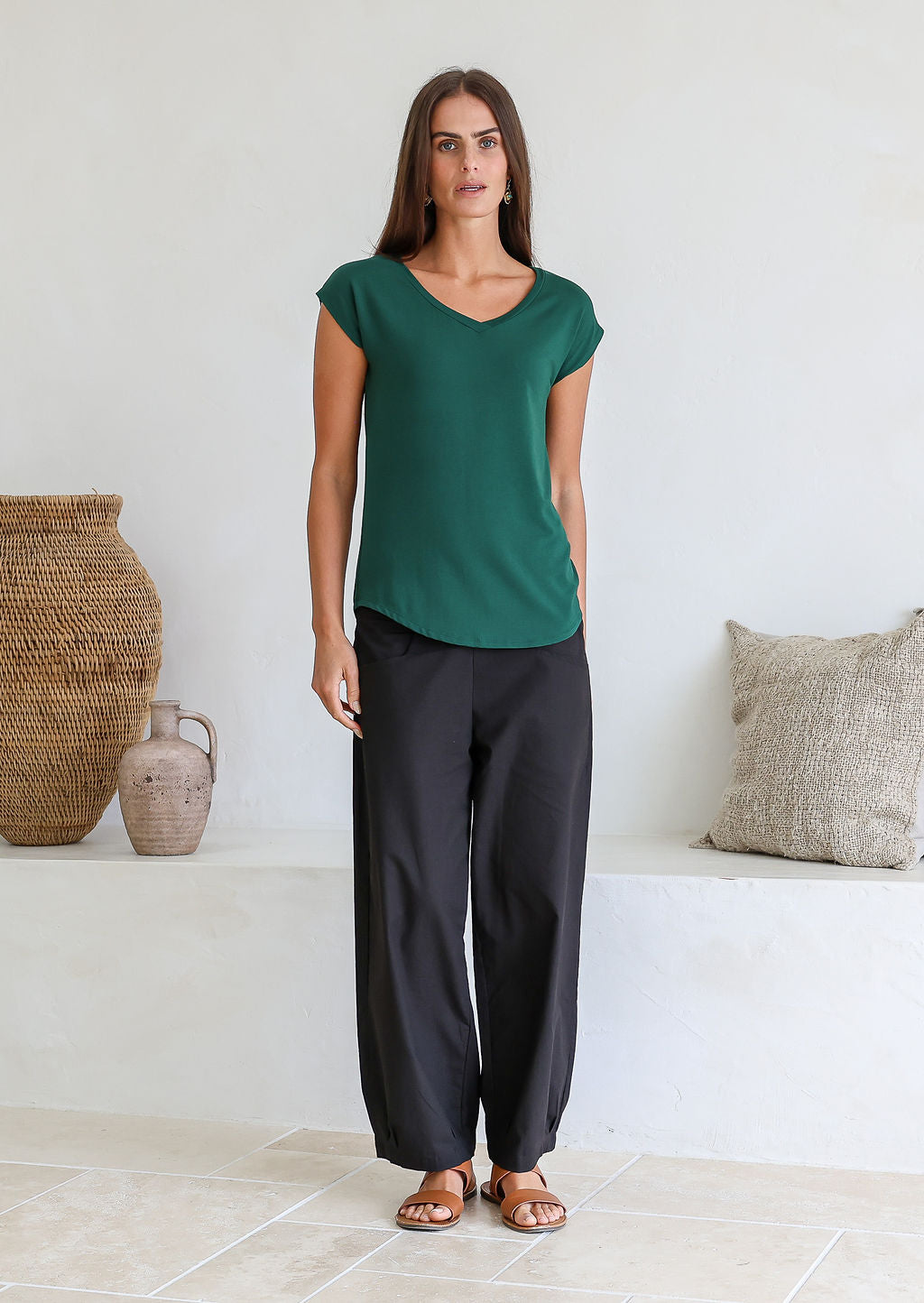Woman wearing a 100% rayon foresr green V-neck top and black pants standing in a minimalistic room with white wall