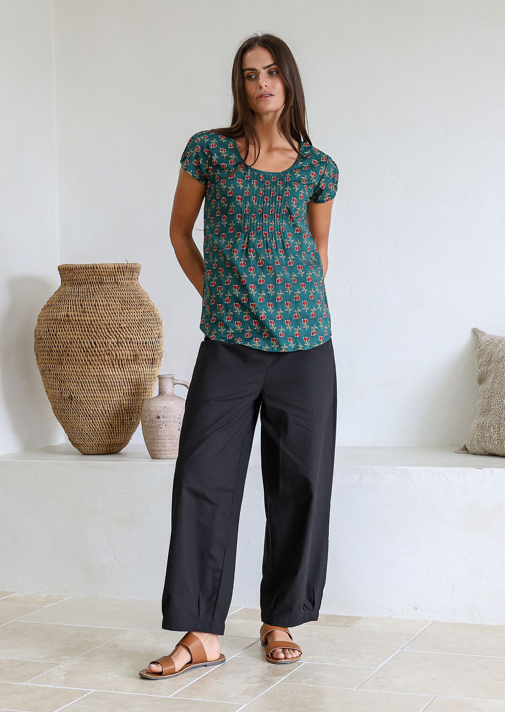 A woman wearing a green, floral-print, 100% cotton top with a U-shaped neckline and cap sleeves, paired with plain black pants.