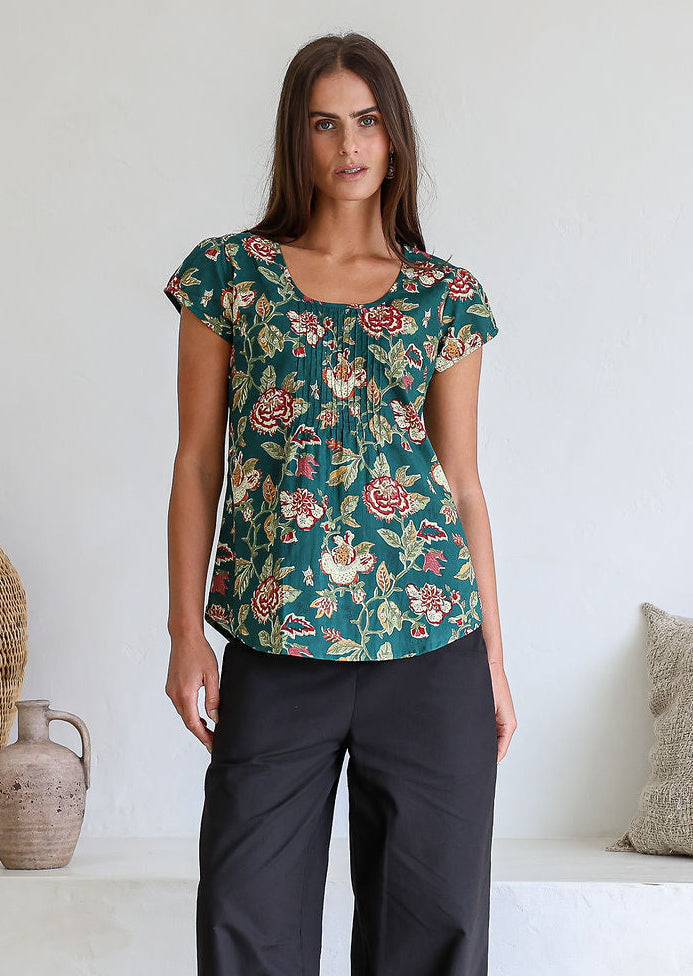 Woman wears bottle green based floral print 100% cotton cap sleeve top with pin tuck detail across the bust and a scoop neckline