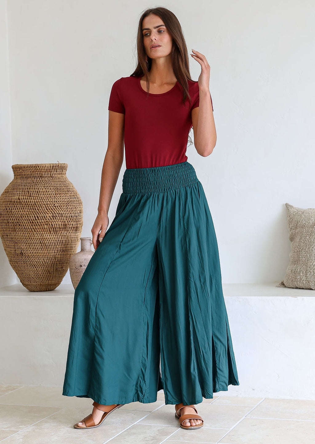 Woman wearing a maroon top and teal wide leg pants standing in a light filled room