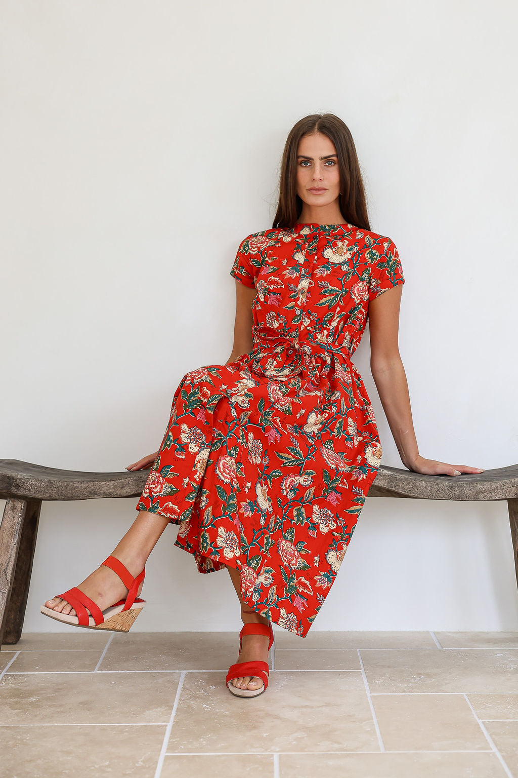 Woman in a red based floral print 100% cotton dress sitting on a wooden bench against a white wall.