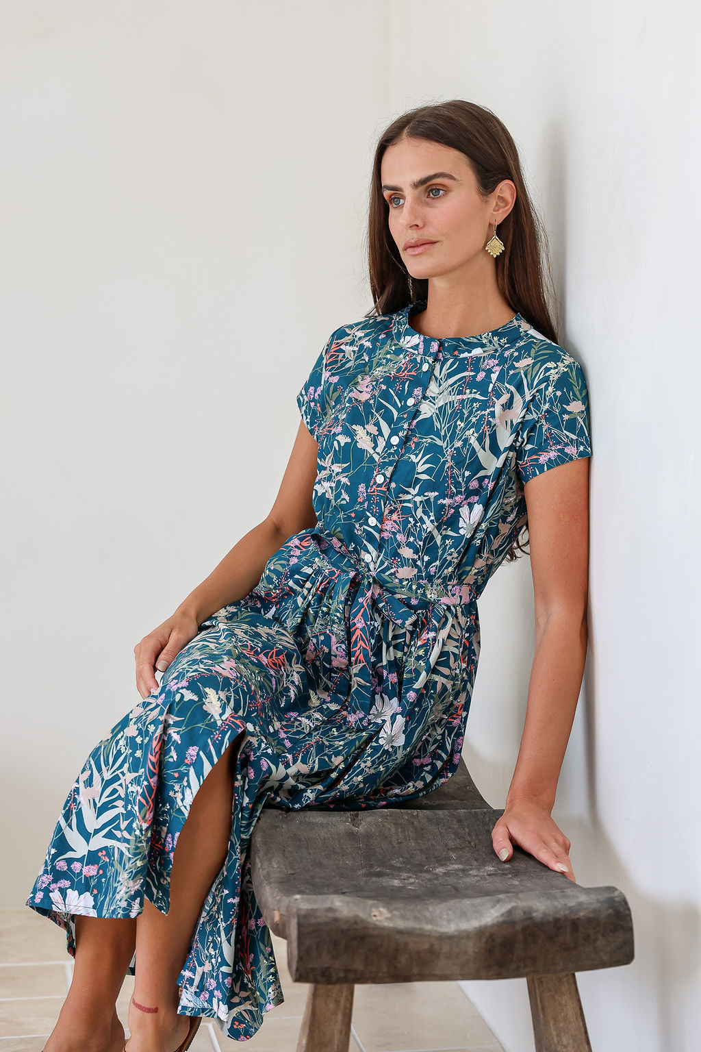 woman sitting on a wooden bench wearing teal based floral printed cotton button through cotton dress with hidden side pockets, short sleeves and a detachable waist tie.