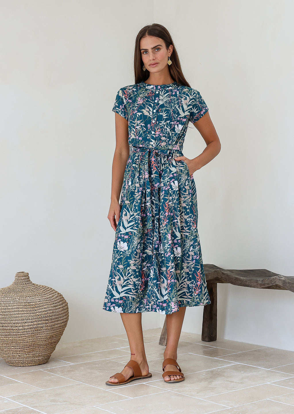 Woman wears 100% cotton button through midi length dress with short sleeves and pockets in floral print on a teal base.