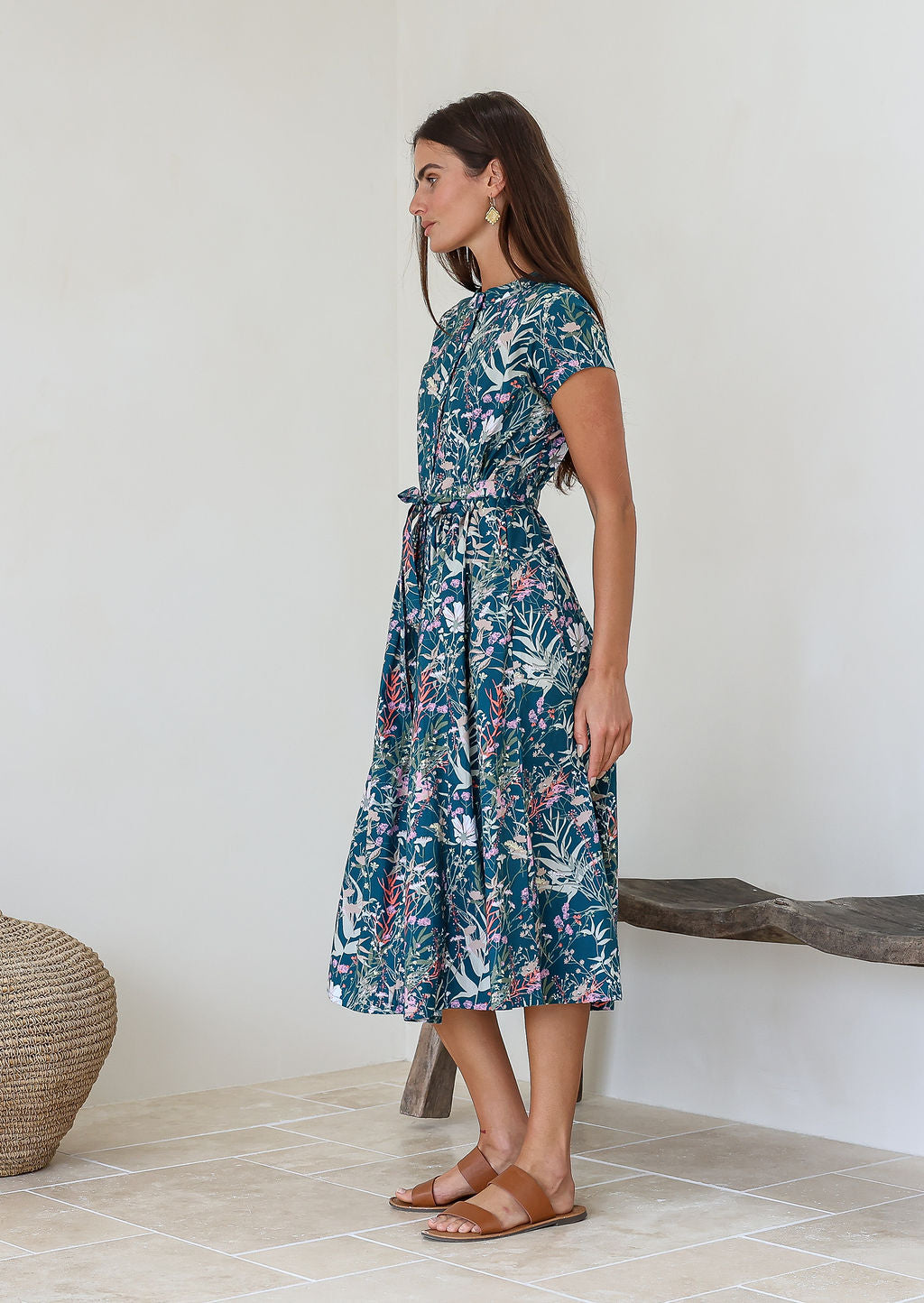 Woman wears teal based floral printed cotton button through cotton dress with hidden side pockets, short sleeves and a detachable waist tie.