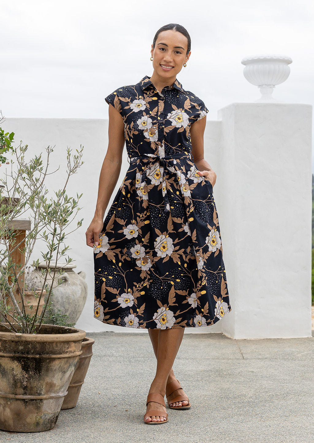 Woman wearing a shirt style cotton midi length dress with buttoned bodice, pockets, collar and cap sleeves