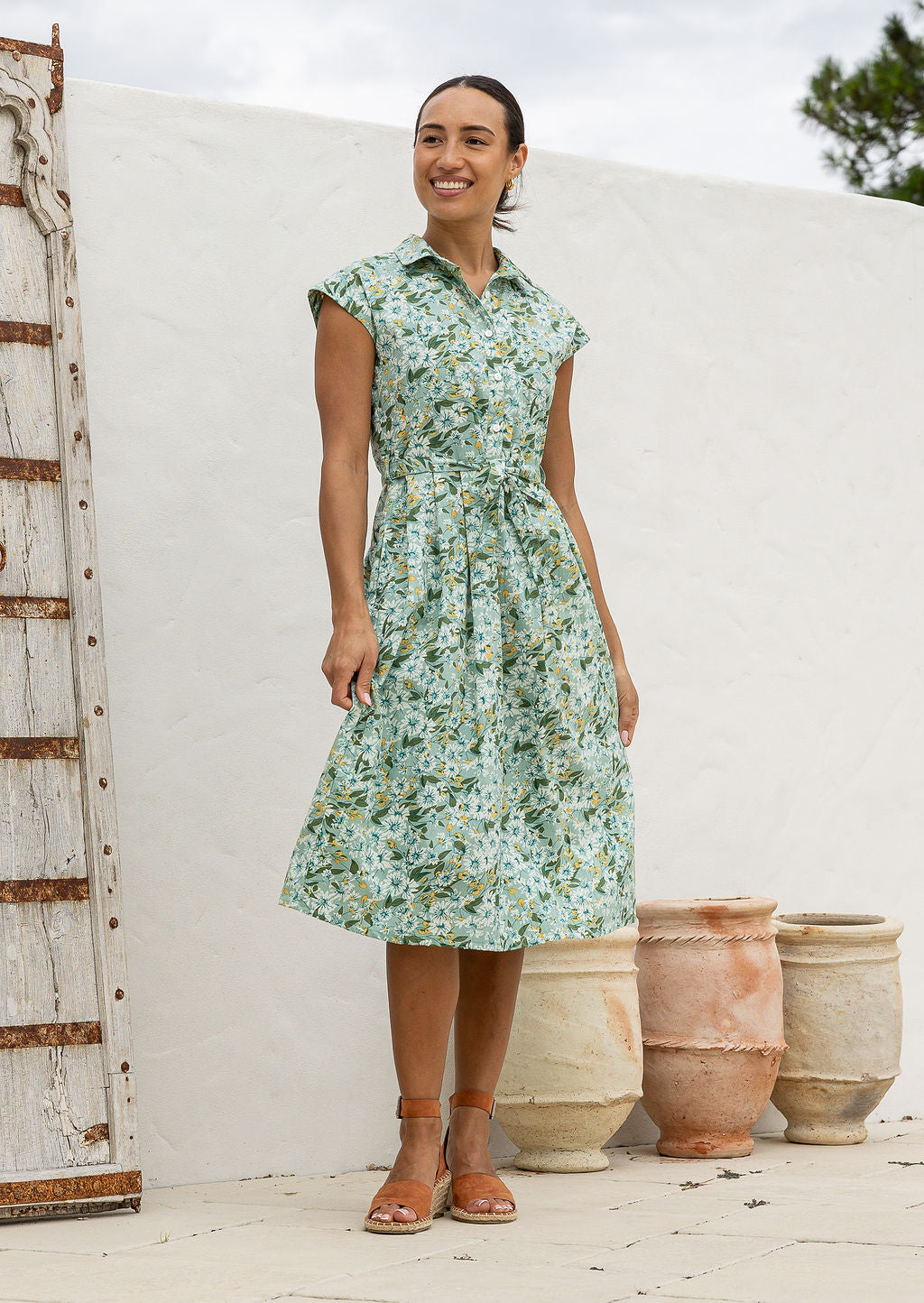Woman wearing a midi length sage green based floral cotton dress with buttoned bodice, standing in front of a white wall with rustic elements.