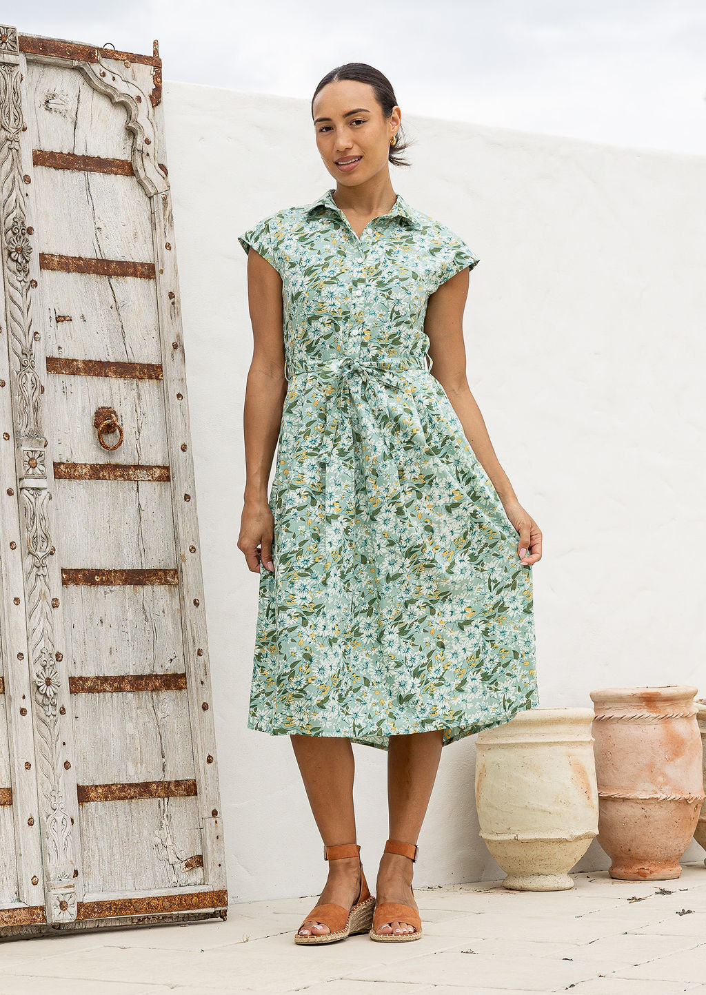 Model stands with wearing a midi length floral printed shirt style cotton dress with cap sleeves, buttoned bodice and a removable waist tie