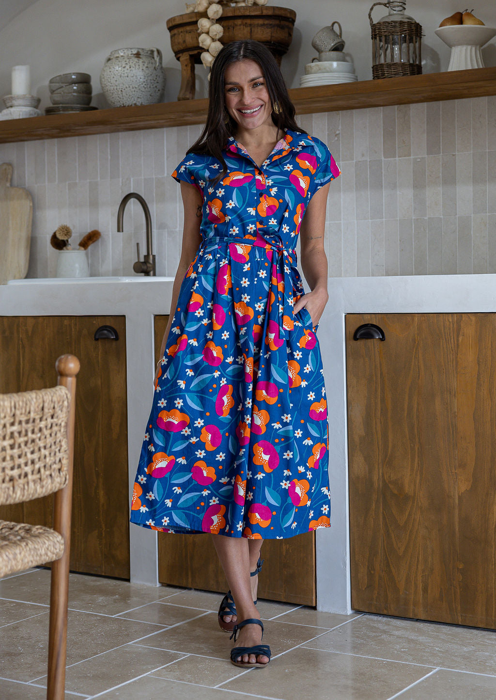 Model stands with wearing a midi length floral printed shirt style cotton dress with cap sleeves, buttoned bodice and a removable waist tie