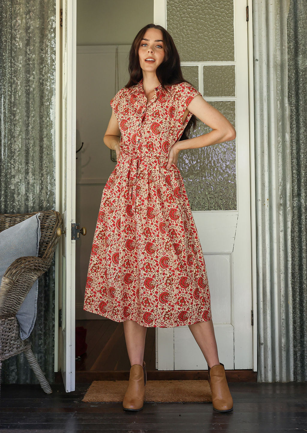 Model stands with wearing a midi length floral printed shirt style cotton dress with cap sleeves, buttoned bodice and a removable waist tie
