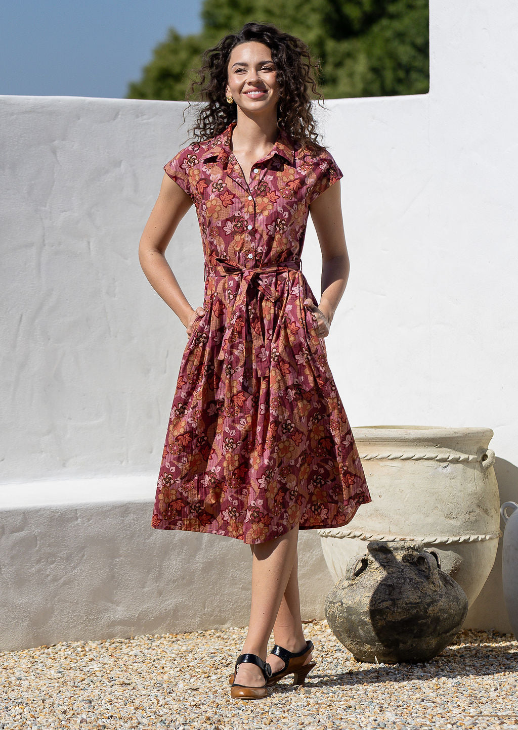 Model stands with both hands in pocket wearing a midi length floral printed shirt style cotton dress with cap sleeves, buttoned bodice and a removable waist tie