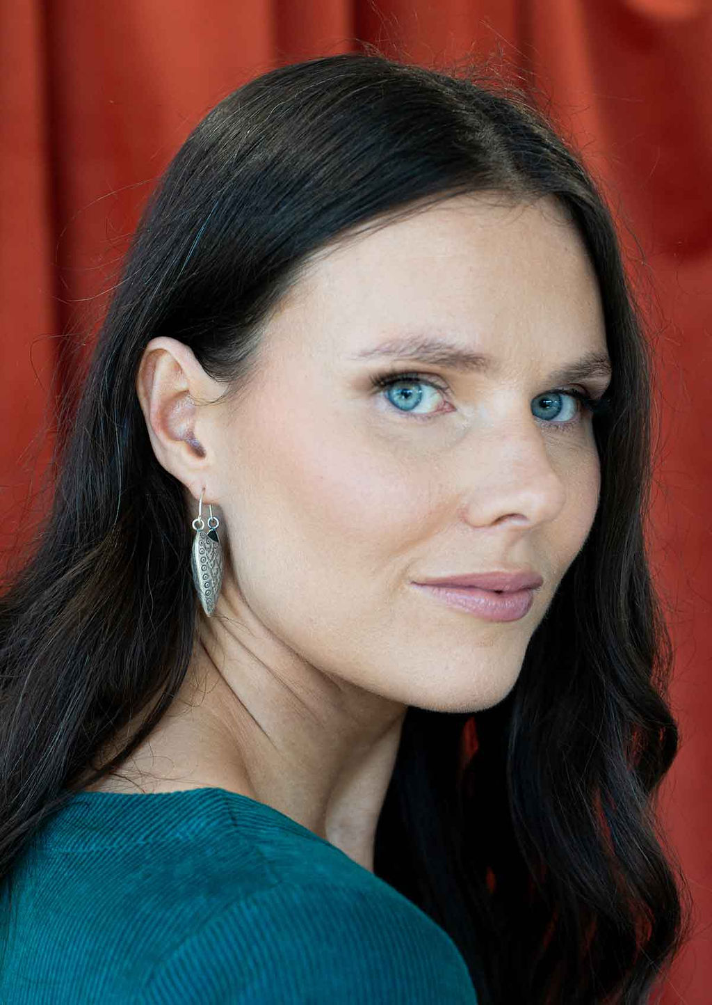 Woman with dark hair and blue eyes wearing Hill Tribe Silver earrings against a red curtain background