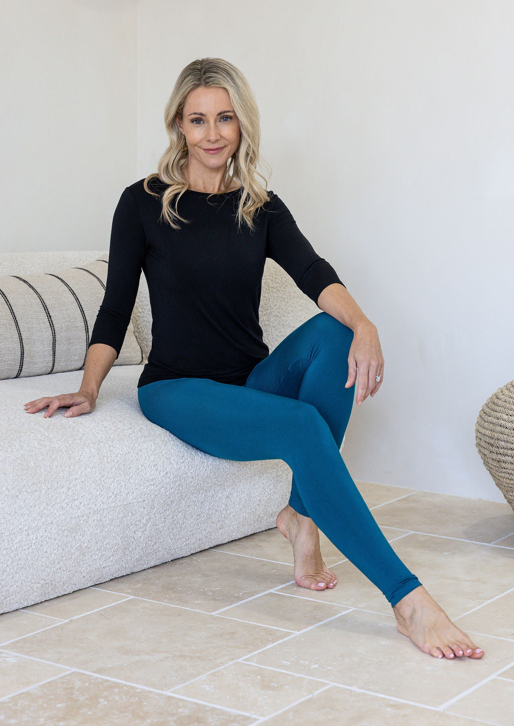 A woman sits on a textured white sofa wearing a black long-sleeve top and teal blue leggings.