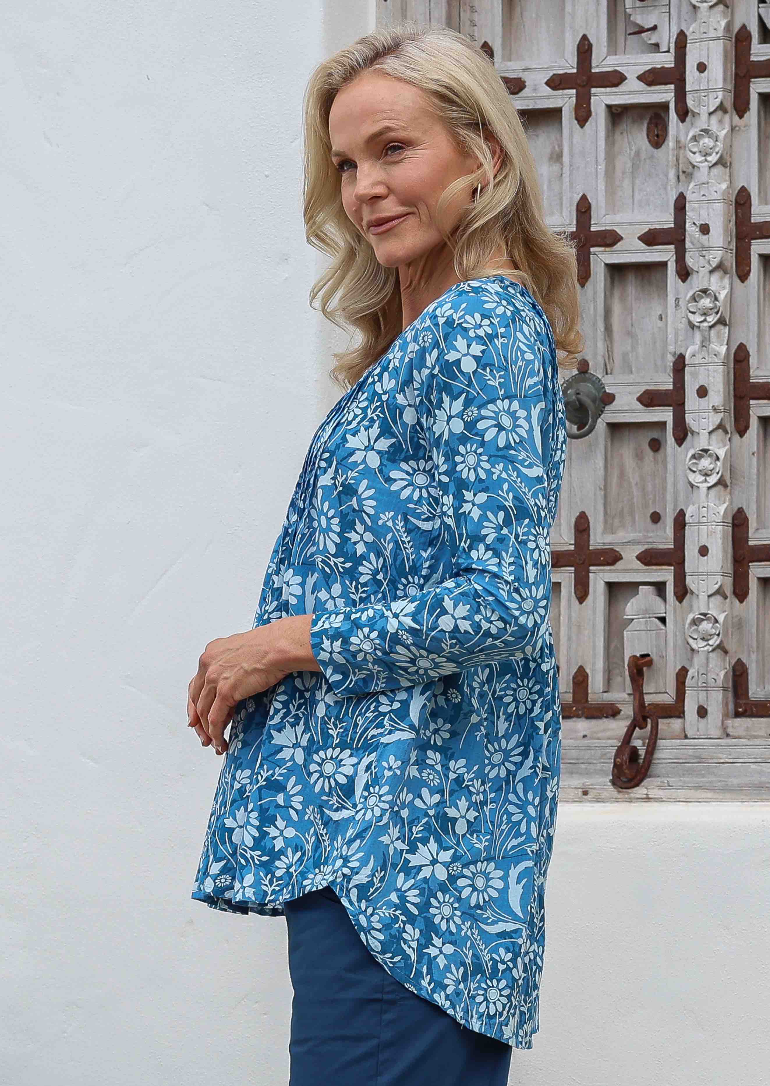 Woman wearing a blue floral button through cotton top with long sleeves, pleats at the front and back, and a longer hem at the back.