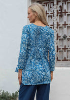 Woman standing in front a wooden window wears 100% cotton pleated front and back detail full sleeve top with V neckline and A line shape.